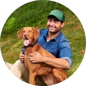man wearing green hat and blue shirt holding vizsla pointer looking off in distance